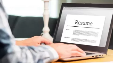Person writing a resume on a laptop in a home setting.