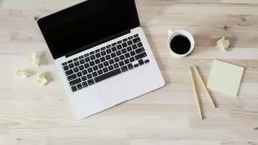 Open laptop on wooden desk with coffee, pencils, sticky notes, and crumpled paper.